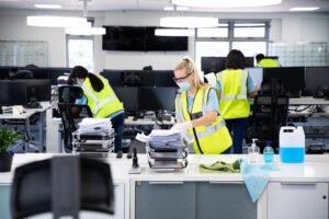 caucasian woman and colleagues wearing hi vis vests, gloves, safety glasses and face masks sanitizing an office using disinfectant. hygiene in workplace during coronavirus covid 19 pandemic.
